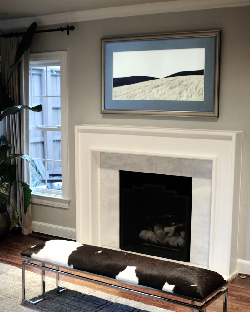 A white marble fireplace under a framed landscape print, a cowhide bench in front