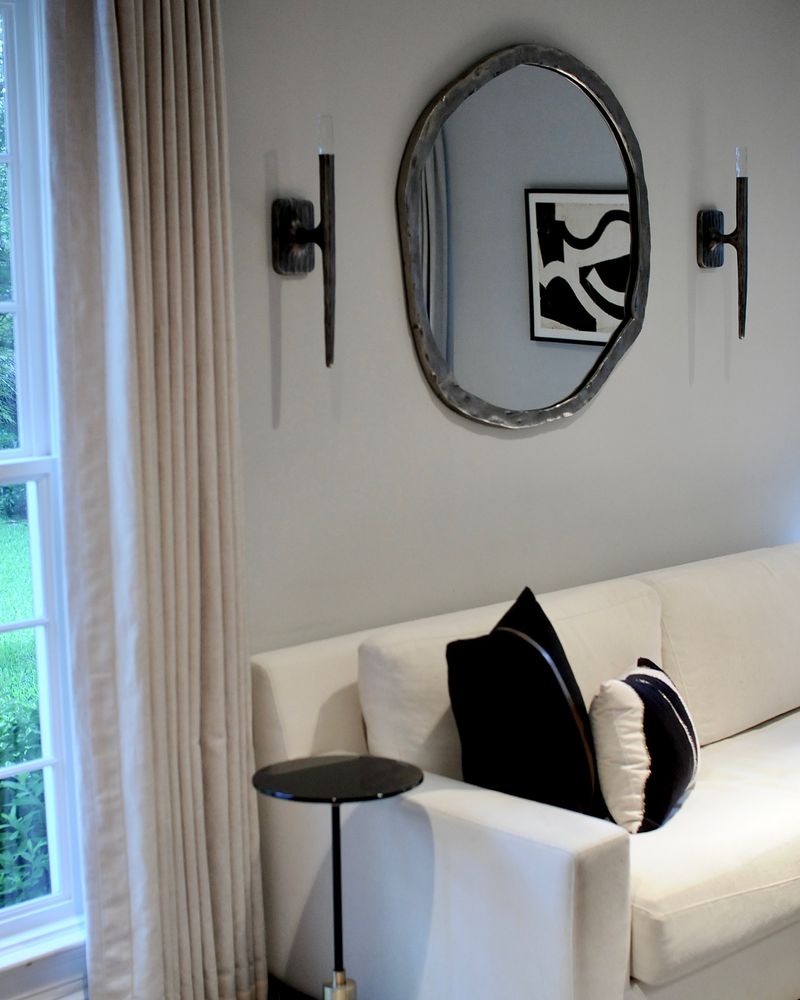 A sculptural oval mirror between two torch sconces above a cream sofa