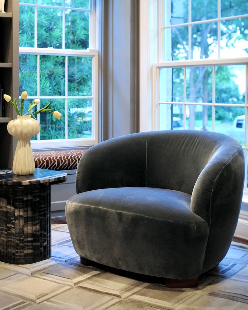 A grey velvet swivel chair and a marble side table with tulips beside tall windows